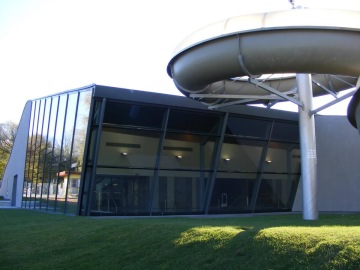 Moderní budova s prosklenou fasádou a tobogánem, trávník vpředu.