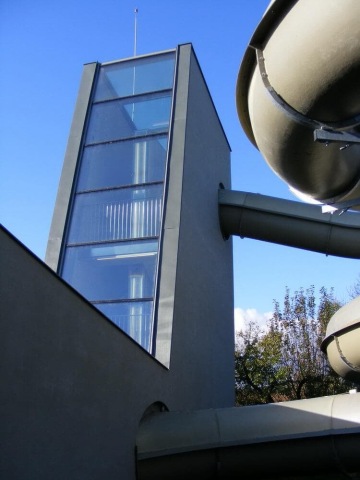 Skleněná věž s šedými tobogány za slunečného dne.