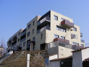 Moderní bytový dům Mazanka s balkony, stojící na vyvýšeném terénu, proti modré obloze.