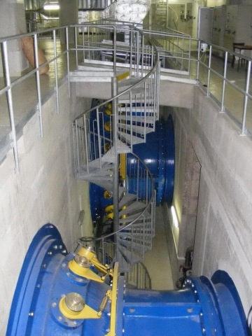 Schodiště ve tvaru spirály vedoucí kolem velkého modrého potrubí v průmyslové hale.