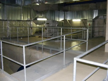 Průmyslový ventilátor a potrubí ve strojovně s kovovými zábradlími.