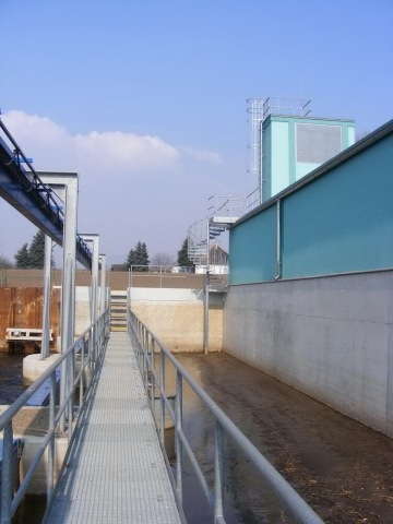 Ocelová lávka vedoucí k průmyslovému objektu s modrými stěnami za jasného dne.