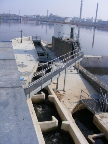 Jezová přehrada s kovovými zábradlími a průtokem vody, tovární komíny v pozadí.