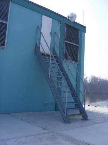 Exteriérové schodiště vedoucí k zavřeným dveřím na modré budově u řeky.