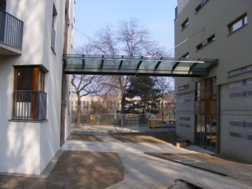 Skleněný průchod mezi dvěma moderními budovami Pod Tyršovým vrchem, s venkovními balkony a bezlistými stromy v pozadí.