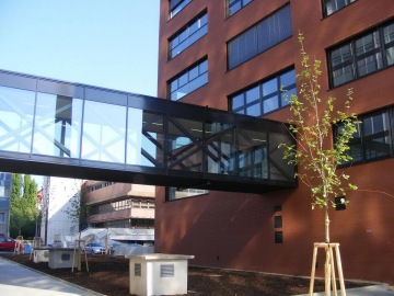 Prosklená lávka spojující cihlovou budovu ČVUT Praha Fakulta stavební s dalším objektem, s mladým stromkem před budovou.