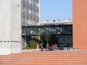 Chodník lemovaný cihlovými zídkami, chodec vedle zaparkovaných motorek, moderní skleněná budova ČVUT Praha Fakulta stavební s přemostěním.