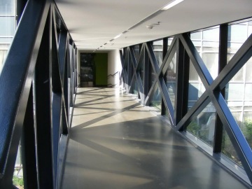 Prosklená chodba ČVUT Praha Fakulta stavební s kovovou konstrukcí a stíny na podlaze během slunečného dne.