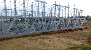 Ocelová konstrukce před rozvodnou stanicí s elektrickými vedeními a stožáry v pozadí.