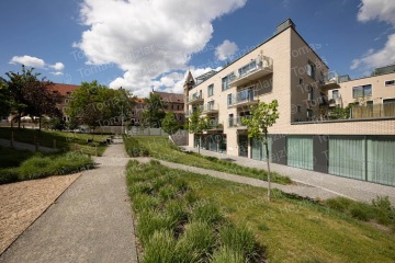 Moderní obytný dům s prosklenými balkony obklopený zeleným parkem pod slunečnou oblohou.