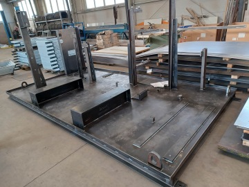 Ocelová konstrukce na dílenské podlaze obklopená kovovými a dřevěnými deskami v průmyslovém prostředí.