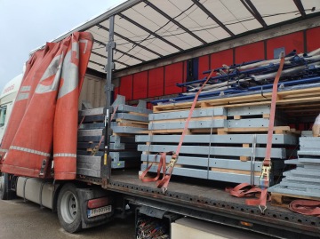 Nákladní auto s otevřenou plachtou, přepravující kovové konstrukce naložené na dřevěných paletách, zajištěné červenými popruhy.