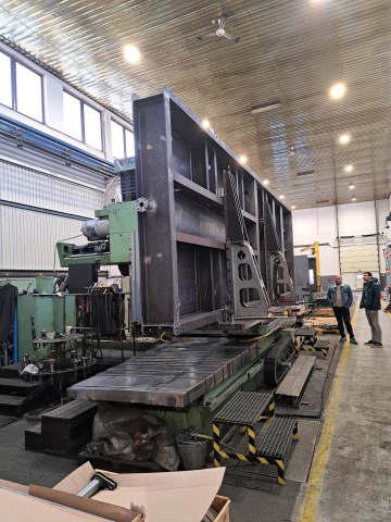 Velká kovová konstrukce na obráběcím stroji v tovární hale, dva muži diskutují.