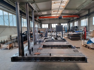 Výrobní hala s kovovou konstrukcí na podlaze a vysokými sloupy, pracovníci v pozadí.