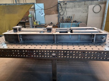 Pracovní stůl s kovovou konstrukcí v dílně, pozadí s nářadím a zásobníky.