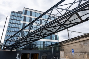 Moderní ocelový most spojující kancelářskou budovu, nad dopravní infrastrukturou pod zataženou oblohou.