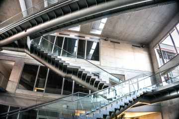 Moderní interiér s křížícími se schodišti a skleněnými zábradlími, světlé betonové stěny.