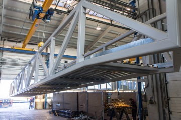 Velká ocelová konstrukce zavěšená na jeřábu v tovární hale, kde probíhá svařování s jiskrami.