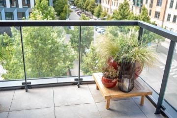 Balkon Marina Nova s čirým sklem, květináče s travinami na dřevěném stolku, zelené stromy a budovy v pozadí.