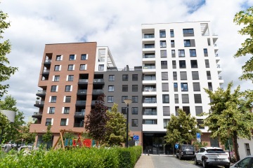 Moderní bytový dům Marina Nova s červenou, šedou a bílou fasádou, auta zaparkovaná před budovou.