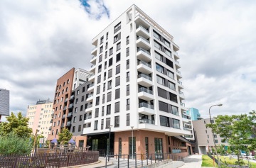 Moderní bytový dům Marina Nova s bílou fasádou a velkými okny, obklopený městskou zelení pod zamračenou oblohou.