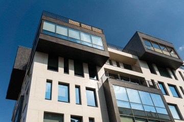 Moderní bytový dům s velkými okny a balkony, modrá obloha v pozadí.