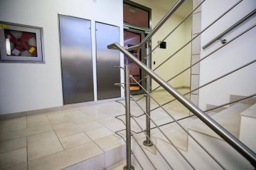 Schodiště s nerezovým zábradlím vedoucí do chodby s dveřmi a hasicí stanicí na zdi.