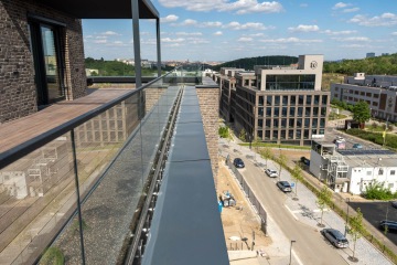 Balkon projektu Nová Waltrovka s proskleným zábradlím, výhled na moderní kancelářské budovy a ulici, slunečný den.