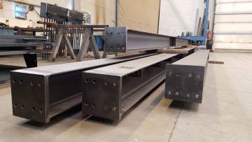 Ocelové nosníky s otvory, ležící na podlaze továrny.