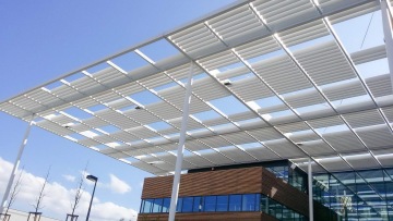 Moderní budova s prosklenou fasádou a bílou ocelovou konstrukcí pod modrou oblohou.