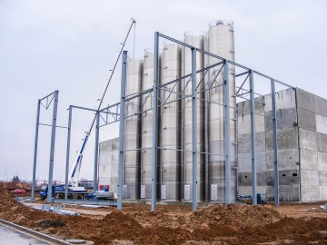 Ocelová konstrukce rozestavěné průmyslové budovy s jeřábem a stříbrnými silami na šedém podkladu.