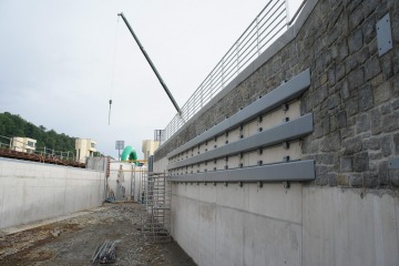 Stavební oblast s jeřábem, betonovou zdí a ocelovými nosníky, okolní les v pozadí.