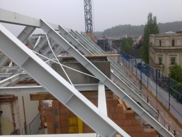 Ocelová konstrukce střechy na rozestavěné budově s kovovými trámy, cihelnými zdmi a stavebním jeřábem v pozadí.