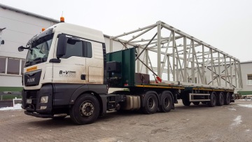 Kamion převážející velkou ocelovou konstrukci na přívěsu, zaparkovaný na dlážděném dvoře u průmyslové budovy.