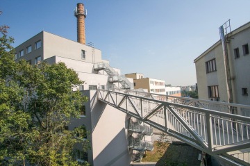 Budova s externími schodišti a komínem na pozadí, s ocelovým mostem v popředí pod modrou oblohou.
