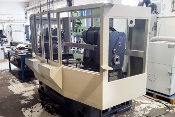 Průmyslový stroj v dílně s nedokončenou konstrukcí a kovovými součástkami.