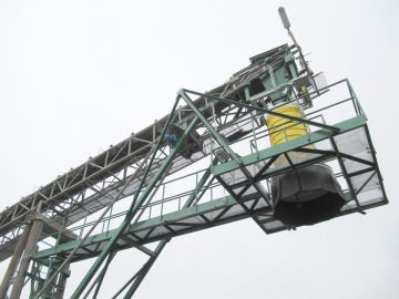 Průmyslový dopravník s ocelovou konstrukcí a žlutým válcem proti šedé obloze.