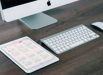 Monitor, klávesnice a tablet s otevřeným kalendářem na dřevěném stole.