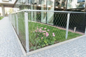 Kovové pletivo kolem záhonu s růžovými květinami u moderní budovy.