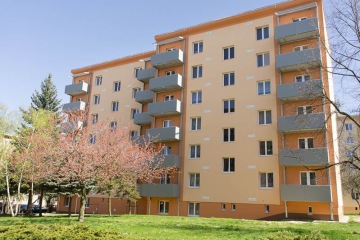 Žlutá bytovka se slunečnými lodžiemi obklopená zelení a stromy v slunečný den.