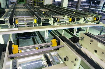 Strojní zařízení s kovovými kolejnicemi a žlutými plastovými součástmi v tovární hale.