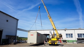 Žlutý jeřáb zvedající bílý kontejner na nákladní rampu před průmyslovou halou pod modrou oblohou.