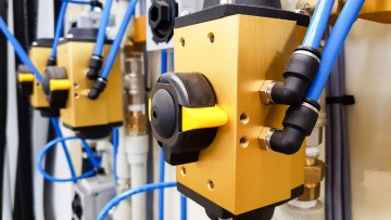 Zlatý ventilační ventil s černou rukojetí, připojený k modrým plastovým trubkám.