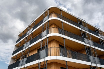 Moderní bytový dům Timber s dřevěným obložením a černým zábradlím na pozadí zamračené oblohy.