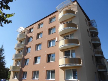 Šestipatrový bytový dům se zaoblenými balkony a satelitní anténou za slunečného dne.