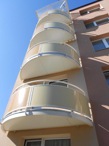 Pohled na moderní zaoblené balkony čtyřpodlažní budovy proti modré obloze.