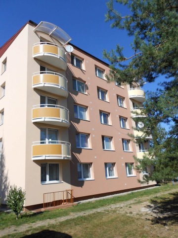 Pětipatrová bytová budova se zaoblenými balkony a stromem v popředí pod modrou oblohou.