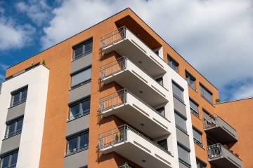 Moderní bytový dům s balkony a oranžovou fasádou na pozadí modré oblohy.