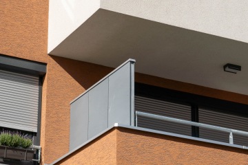 Oranžová fasáda budovy s šedým zábradlím na balkóně a zavřenými roletami.