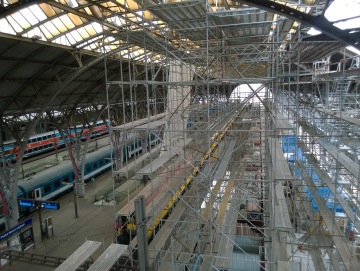 Železniční nádraží obklopené lešením s vlaky na nástupištích, rekonstrukce probíhá.